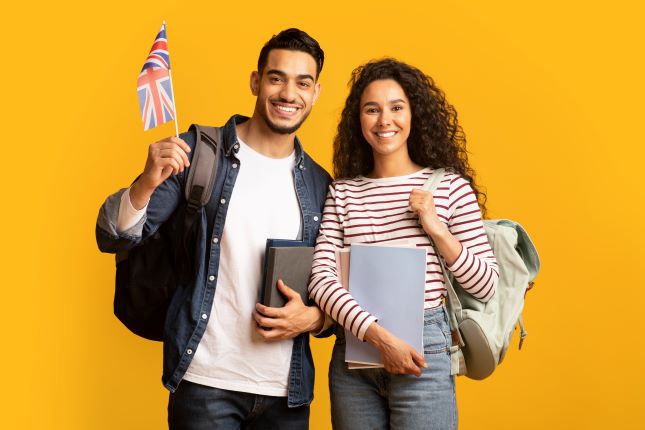 Para ludzi, prawdopodobnie studentów - młoda kobieta i młody mężczyzna, stoją obok siebie i uśmiechają się. Mężczyzna trzyma w ręku flagę Wielkiej Brytanii.